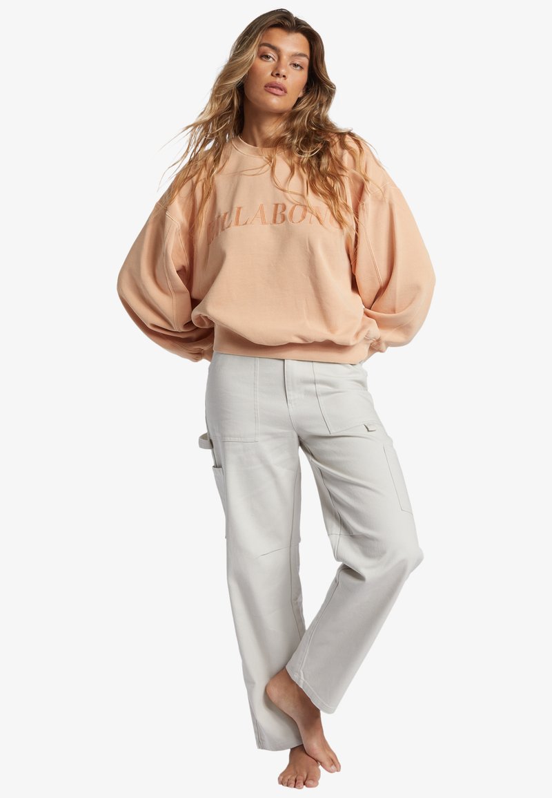 Sudadera de gran tamaño color durazno con el texto "BILLABONG"; combinada con pantalones cargo rectos de color gris claro que presentan bolsillos laterales y una textura suave.