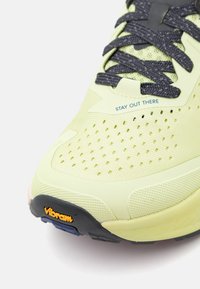 Scarpa da corsa verde chiaro con upper testurizzato, lacci grigi e marchio Vibram. Presenta perforazioni per la traspirabilità e design della suola robusto.