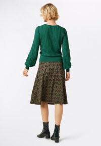 Grüner strukturierter Pullover mit puffigen Ärmeln, kombiniert mit einem knielangen, blumigen Rock in dunklen Tönen, akzentuiert durch glänzende schwarze Ankle Boots.