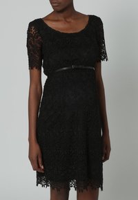 Czarna koronkowa sukienka z krótkimi rękawami, z misternym wzorem kwiatowym. Posiada wąski czarny pasek w talii. Spódnica z falbaniastym dołem.