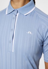 Polo shirt azzurro chiaro con texture a righe verticali, dettagli bianchi sul colletto e un logo sul lato sinistro del petto. Tre bottoni frontali visibili.