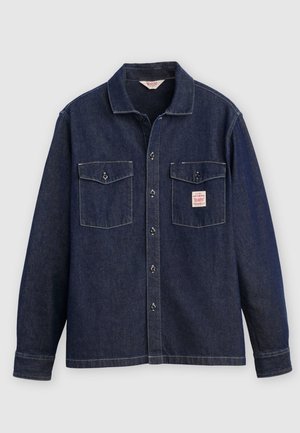 Cămașă din denim închis la culoare, cu guler, două buzunare pe piept și nasturi în față. Prezintă cusături contrastante și o etichetă pe buzunarul din stânga.