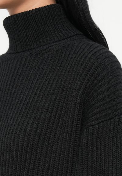 Gros plan sur une personne portant un pull à col roulé côtelé noir, avec de longs cheveux noirs visibles sur l'épaule.