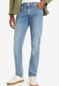 Vaqueros de mezclilla azul claro con un corte recto, diseño de cinco bolsillos y un leve desgaste. Combinados con zapatillas blancas y una camisa verde.