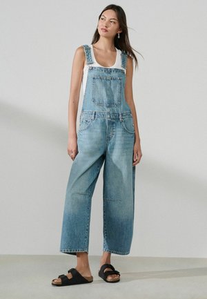 Frau steht, trägt hellblaue Jeans-Latzhose, weißes ärmelloses Top und schwarze Sandalen vor einem schlichten hellgrauen Hintergrund.