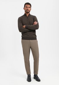 Bruine ritsvest met een textuurfinish, gecombineerd met beige broek en zwarte enkellaarsjes. Model staat met gekruiste armen tegen een witte achtergrond.
