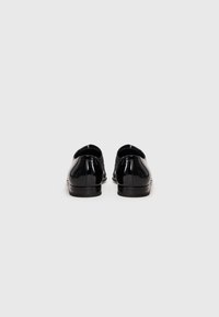 Un par de zapatos de vestir negros brillantes mostrados desde la parte trasera sobre un fondo blanco liso.