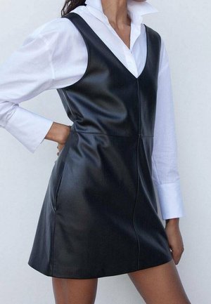 Femme portant une robe sans manches en cuir noir par-dessus une chemise blanche à manches longues avec col, main posée sur la hanche.