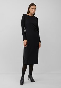 Frau in einem langärmligen schwarzen Midikleid mit schwarzen Strumpfhosen und schwarzen Stiefeletten, die vor einem schlichten grauen Hintergrund steht.