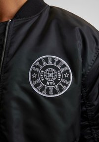 Veste bomber noire en nylon lisse, avec un logo circulaire brodé en blanc comprenant un texte et un motif de globe sur le côté gauche de la poitrine.