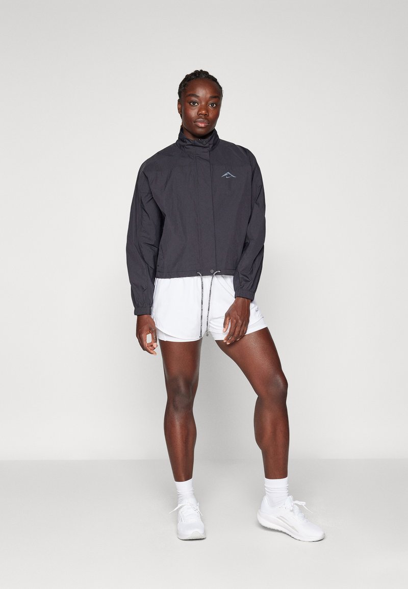 Frau, die in weißen Sportshorts, weißen Turnschuhen und einer schwarzen Reißverschlussjacke mit Kordelzügen vor einem schlichten hellen Hintergrund steht.