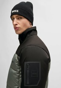 Black knit beanie with "BOSS" logo; black and gray padded jacket with high collar and zip pocket on left arm. Smooth texture and modern design.