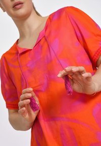 Heldere oranje top met roze tie-dye patroon, met een v-hals, korte mouwen en kwastdetails aan de trekkoorden. Glad katoenstof.