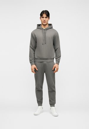 Άνδρας που στέκεται στραμμένος προς τα εμπρός φορώντας γκρι φούτερ με κουκούλα και αντίστοιχα παντελόνια τζόγκινγκ με λευκά αθλητικά παπούτσια σε απλό φόντο.