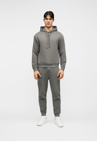 Homme debout face à l'avant, portant un sweat à capuche gris et un pantalon de jogging assorti, avec des baskets blanches, sur un fond uni.