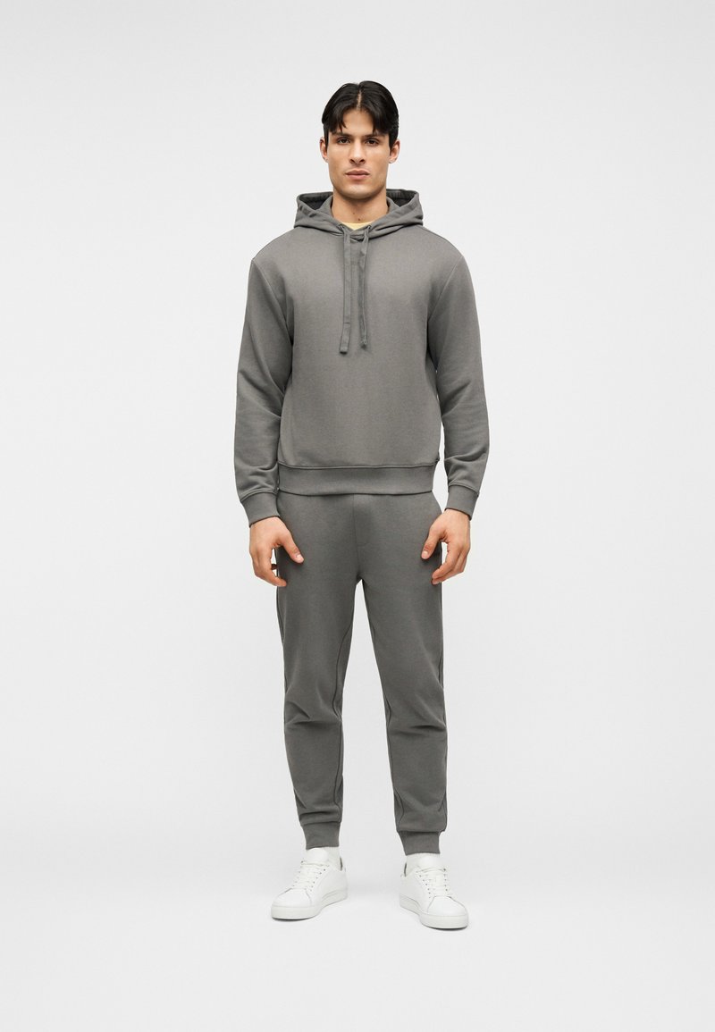 Άνδρας που στέκεται στραμμένος προς τα εμπρός φορώντας γκρι φούτερ με κουκούλα και αντίστοιχα παντελόνια τζόγκινγκ με λευκά αθλητικά παπούτσια σε απλό φόντο.