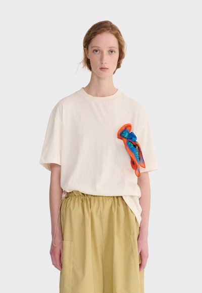 Jeune personne à la peau claire et aux cheveux roux portant un t-shirt ample écru avec une écharpe colorée dans la poche poitrine et un pantalon beige à cordon de serrage.