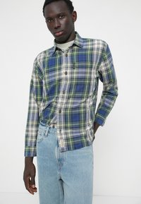Chemise en flanelle écossaise multicolore dans des tons de bleu, vert et jaune, avec des manches longues et une fermeture à boutons ; assortie à un jean en denim bleu clair.