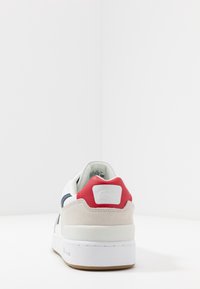 Baskets blanches avec un arrière en daim texturé, un accent rouge sur le talon, une rayure marine et une semelle en caoutchouc blanche. Partie supérieure lisse avec des trous de ventilation.