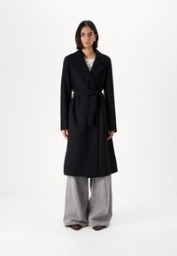 Palton lung negru cu o curea, guler clasic și design cu un singur nasture. Purta peste pantaloni gri cu croi larg, creând o silueta elegantă.