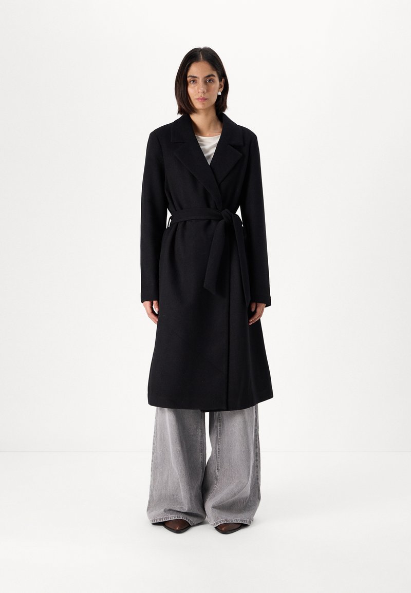 Palton lung negru cu o curea, guler clasic și design cu un singur nasture. Purta peste pantaloni gri cu croi larg, creând o silueta elegantă.