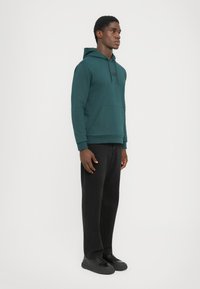 Uomo che indossa un felpa con cappuccio teal, pantaloni neri a gamba dritta e sneakers nere, in piedi contro uno sfondo bianco semplice.