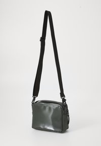 Pravougaona crossbody torba u tamnozelenoj boji, izrađena od sjajnog materijala s glatkom teksturom. Ima crnu podesivu traku i minimalan hardver.
