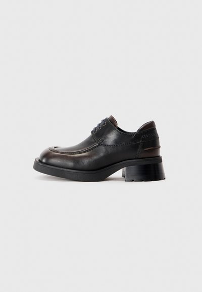 Zapato de cuero negro con punta redondeada, suela de goma gruesa y tacón bajo de bloque. Presenta cordones azules y sutiles acentos marrones en la parte superior.