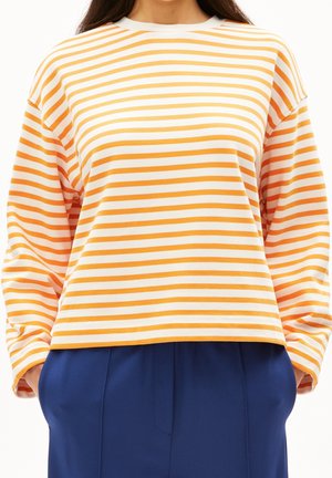 Kvinde iført en langærmet orange og hvidstribet skjorte med blå bukser, stående med hænderne i lommerne mod en hvid baggrund.