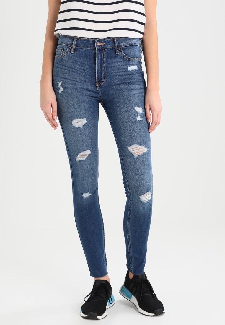 Jeans ajustados en denim de lavado oscuro, con áreas desgastadas y de alta cintura. Combinados con una blusa de rayas en blanco y negro y zapatillas negras.