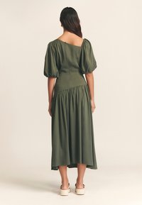 Abito verde oliva con corsetto aderente, maniche a sbuffo e gonna fluida. La parte posteriore presenta una spalla parzialmente scoperta e dettagli arricciati in vita.