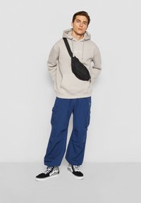 Sweat à capuche beige avec cordons, associé à un pantalon cargo bleu marine avec des poches latérales et des baskets à motif damier noir.
