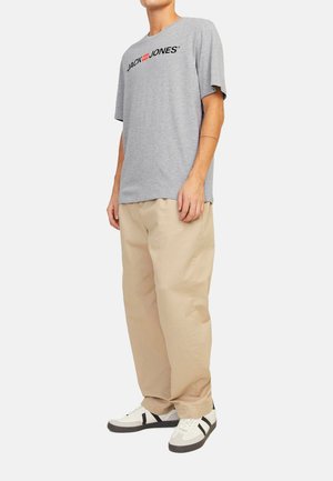 Graues Baumwoll-T-Shirt mit schwarzem Drucklogo, kombiniert mit beigen, lockeren Hosen und weißen Sneakers mit schwarzen Akzenten.