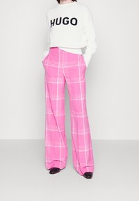 Maglione bianco a coste con logo "HUGO", abbinato a pantaloni a gamba larga a quadri rosa con righe bianche e tasche frontali. Scarpe nere flat.