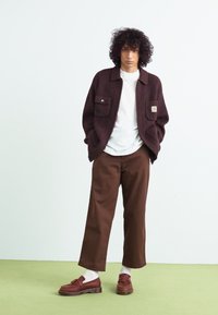Braune Cordjacke mit zwei Taschen, weißem T-Shirt, braunen weit geschnittenen Hosen und braunen Schuhen mit dicker Sohle, vor einem grünen Hintergrund.