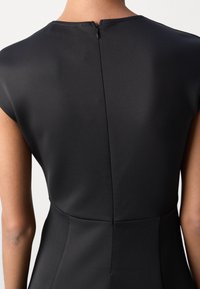 Robe noire ajustée avec manches courtes, un col montant et une fermeture éclair dissimulée dans le dos. Tissu lisse avec des coutures minimales, mettant en valeur une silhouette élégante.