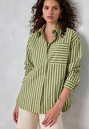 Jonge vrouw met krullend haar die een groen-wit verticaal gestreept overhemd met een horizontaal gestreepte borstzak draagt.
