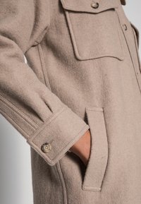 Beige wollen jas met knoopmanchet, klepborstzak en zijzak gedragen aan de linkerarm tegen een effen achtergrond.