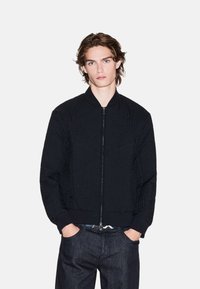 Giacca bomber nera in tessuto testurizzato con un design decorativo, chiusura frontale con zip e polsini e orlo a costine, abbinata a jeans scuri.