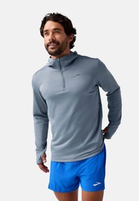 Leichtes, langärmliges graues Laufshirt mit einem halben Reißverschluss und einer Kapuze. Gewebte Textur; Logo-Akzent auf der Brust. Kombiniert mit blauen Shorts.