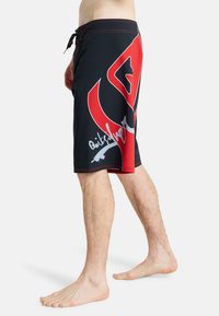 Άνδρας που φοράει μαύρα και κόκκινα κοντά μπανιεράκια με μεγάλο αφηρημένο γραφικό και λευκή γραφή στο πλάι, στέκεται ξυπόλυτος.