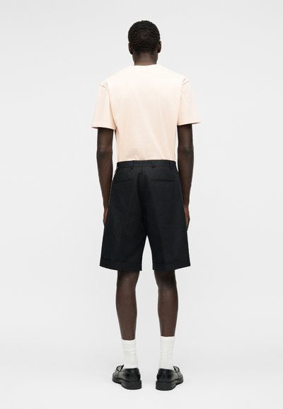 Άνδρας που στέκεται γυρισμένος, φορά ανοιχτό ροδακινί κοντομάνικο πουκάμισο, μαύρα σορτς μέχρι το γόνατο, άσπρες κάλτσες και μαύρα παπούτσια.