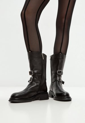Jambes en collants noirs transparents avec une couture verticale, portant des bottes en cuir noires mi-mollet avec des fermetures éclair latérales et des doubles boucles.