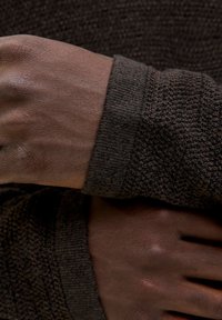 Dunkelbrauner gestrickter Pulloverärmel mit strukturiertem Stoff. Nahaufnahme einer Hand, die dagegen ruht, wobei glatte Haut und feine Details zur Geltung kommen.