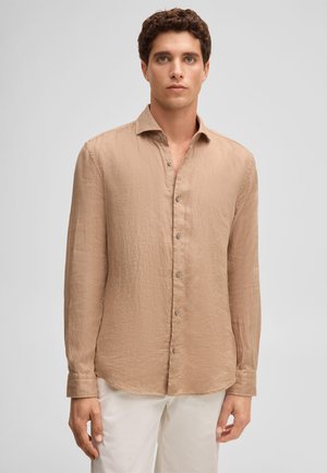 Young man standing wearing a long-sleeve beige button-up shirt and white pants against a plain light gray background.