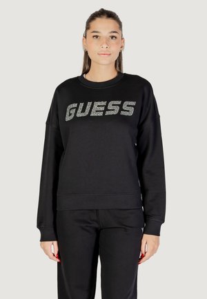 Felpa nera con maniche lunghe e polsini a costine, con un logo "GUESS" luccicante sul petto. Il materiale sembra morbido e strutturato.