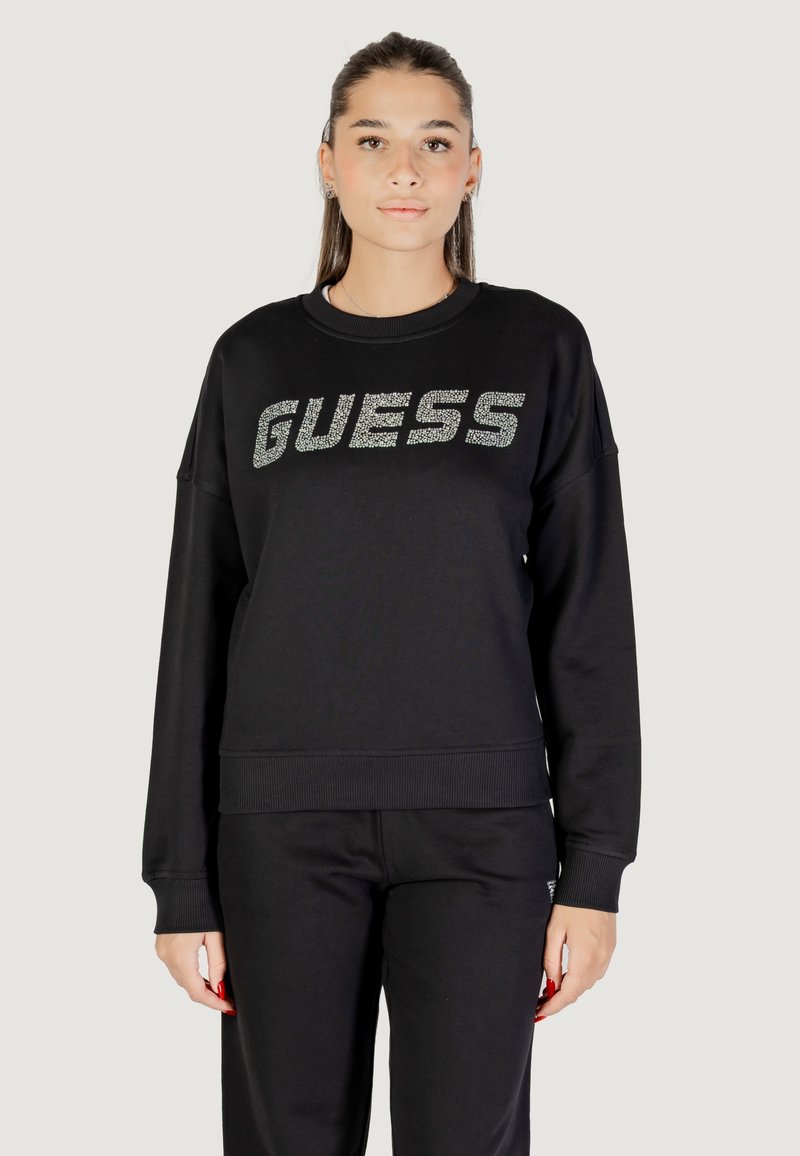 Felpa nera con maniche lunghe e polsini a costine, con un logo "GUESS" luccicante sul petto. Il materiale sembra morbido e strutturato.