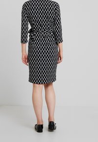 Černo-bíl Dress s diamantovým vzorem, tříčtvrtečními rukávy, zavázaným pasem a délkou ke kolenům. Jsou obuv s černými botami.