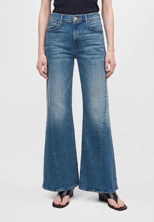 Modèle portant un jean bleu à jambes larges et évasées, un haut noir et des sandales noires sur un fond clair uni.