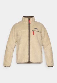 Fleece jakna u krem boji s patentnim zatvaračem sprijeda, smešnim detaljima i crvenim povlačenjem za patentni zatvarač. Ima jedan džep na prsima i dva bočna džepa.